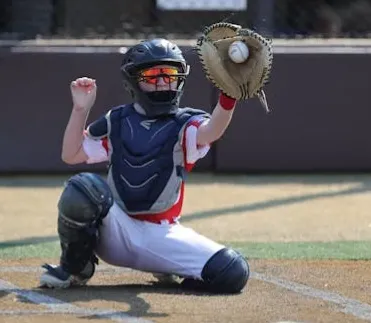 Young baseball catcher in protective gear catching a ball during a game on a sunny day.