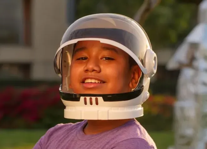 A smiling boy wearing a space helmet outdoors, embodying childhood imagination.