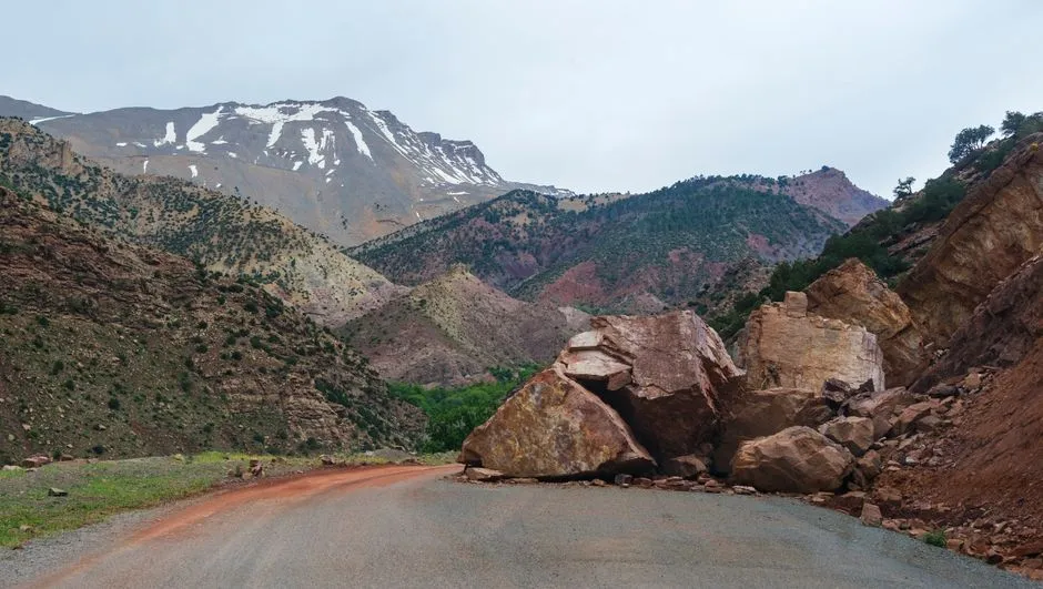 A scenic mountain road blocked by large rocks from a landslide with a snowy peak in the background.