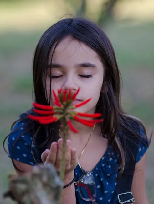 A young girl with brown hair enjoys a moment with a red flower outdoors.