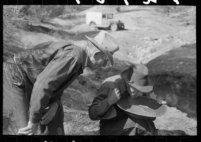 Gold prospector blowing away dirt to find the gold in his pan while a visiting prospector looks on. Pinos Altos, New Mexico