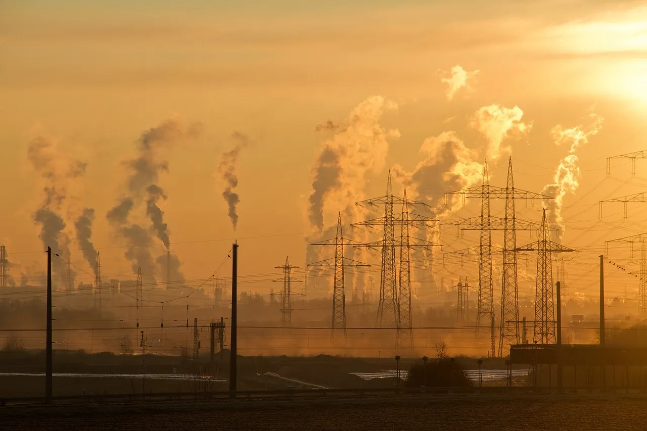 Silhouetted smokestacks and electrical towers against a hazy, golden sunset sky.
